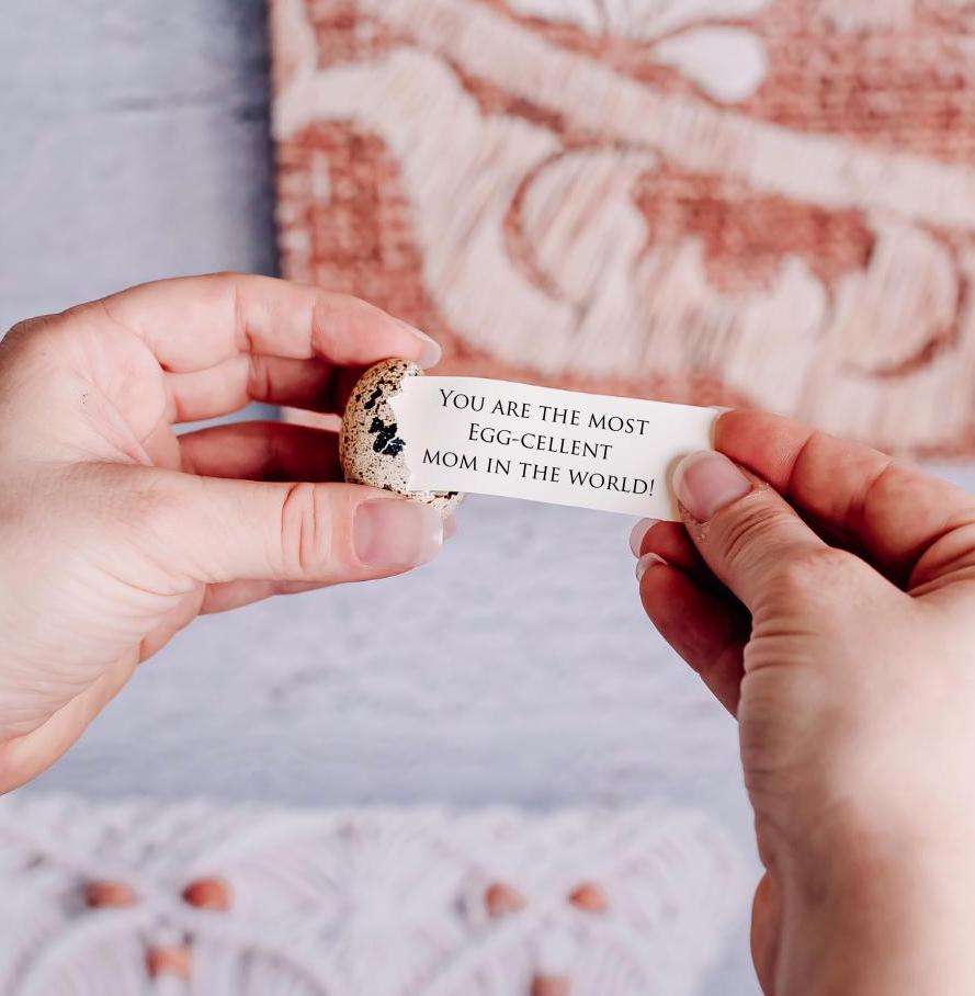 Person holding a small egg with a card that reads 'You are the most egg-cellent mom in the world!' against a blurred background.