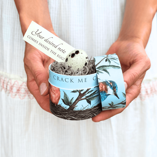 Hand holding a decorative egg with a bird design and a small card inside, against a white background.