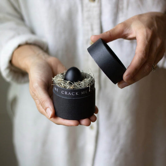 Hands hold a black cylindrical box with the words "crack me" circling around the neck of the box. Inside the box is a nest of spanish moss and a nestled black egg. The egg is a small, quail egg painted black.