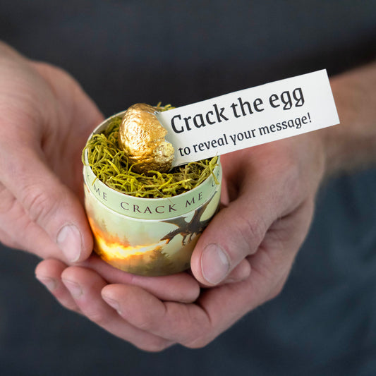 Person holding a small container with a gold egg and a message card, with a blurred background.