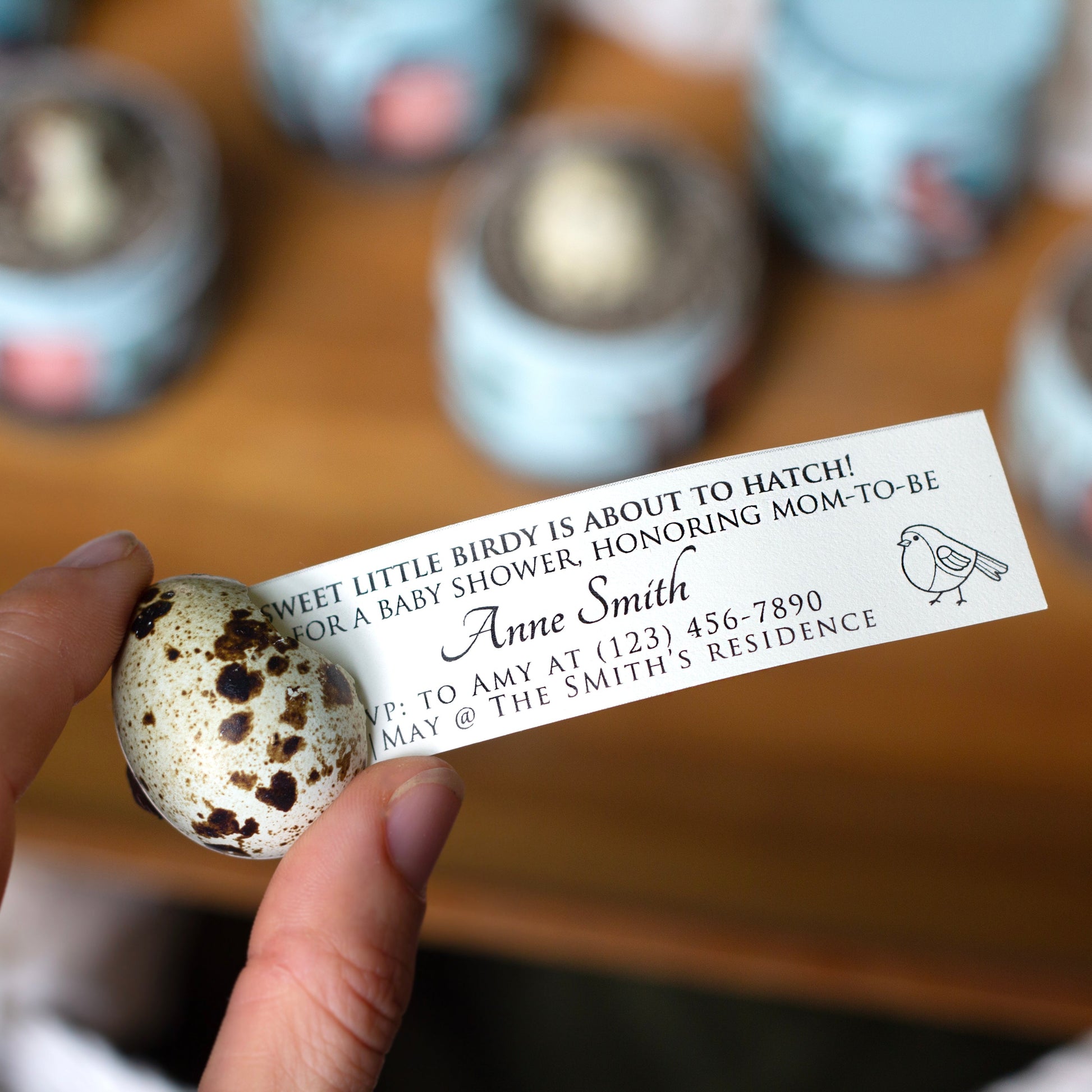 A hand holds a small speckled egg-shaped in front of a blurred wooden surface with several similar  eggs in the background. The card being pulled out of the egg has printed text for a baby shower invitation and a small bird illustration.