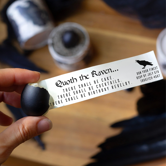A hand holds a black egg with a white paper emerging, printed with gothic text and a raven illustration. The background shows a wooden surface with dark fabric and small containers, suggesting a themed or gothic aesthetic.