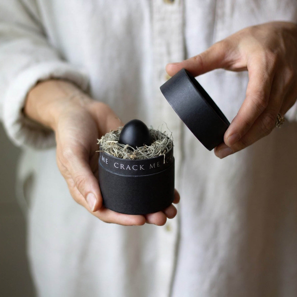 A small black gift box is being opened, within is black egg. The rim of the black box reads "crack me"