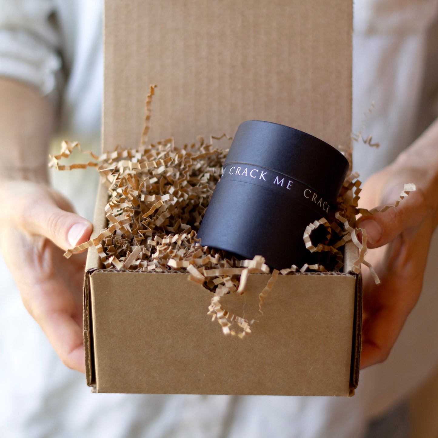 A person holds an open brown cardboard box filled with shredded paper and a black cylindrical box printed with the words 'CRACK ME CRACK ME' in white text.