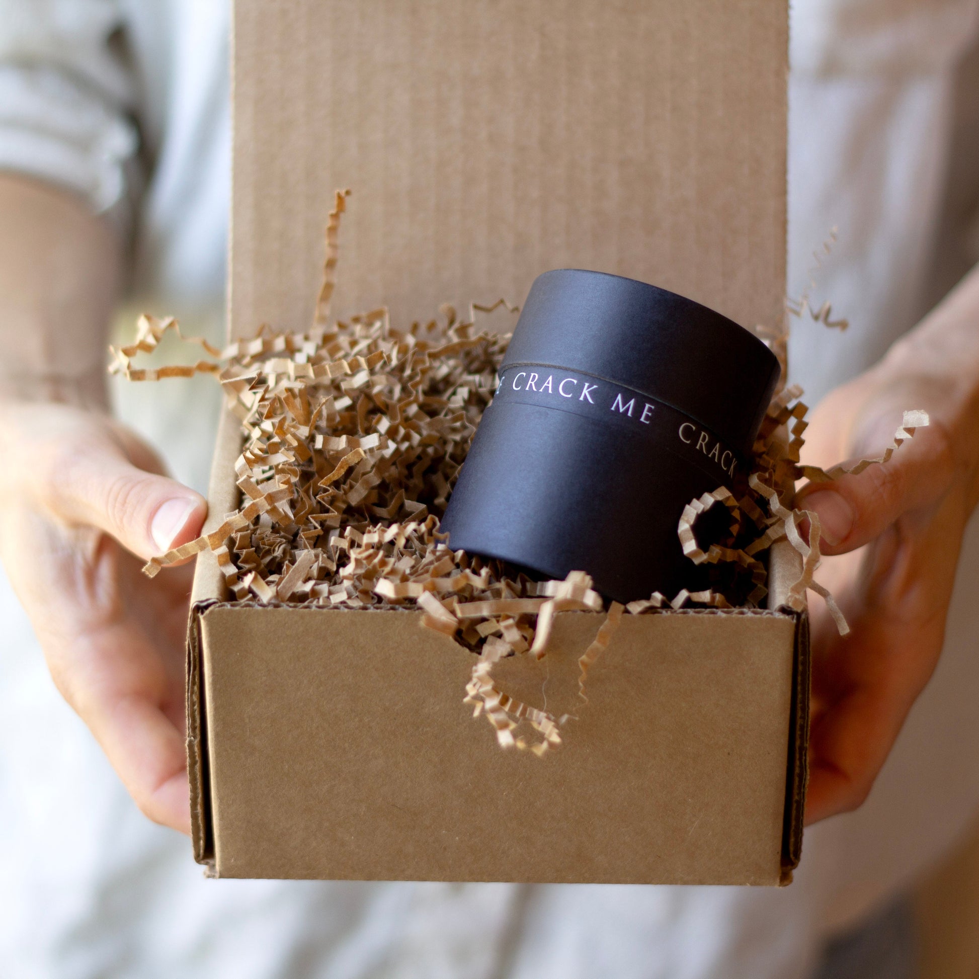 A person holds an open brown cardboard box filled with shredded paper and a black cylindrical box printed with the words 'CRACK ME CRACK ME' in white text.
