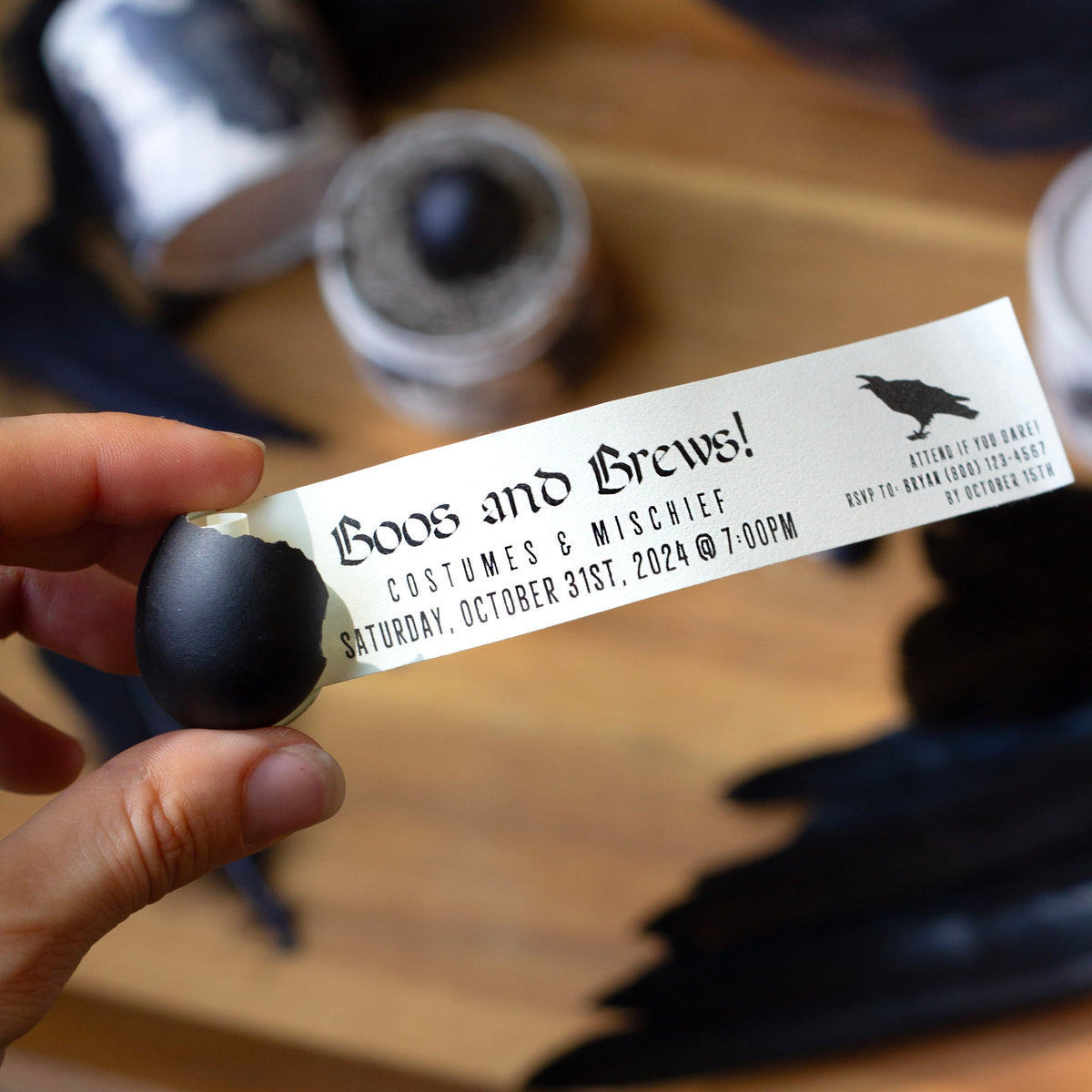 A hand holds a black egg which is cracked and from it emerges a  tag which has a Halloween Party invitation printed on it. Behind is a  wooden table with black feathers and containers in the background