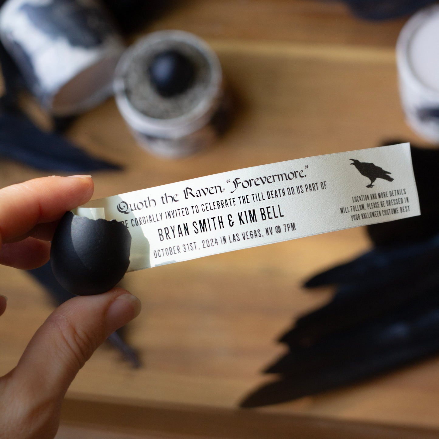 A hand holds a small printed invitation card with black text and a black raven silhouette, surrounded by dark feathers and small glass containers on a wooden surface.