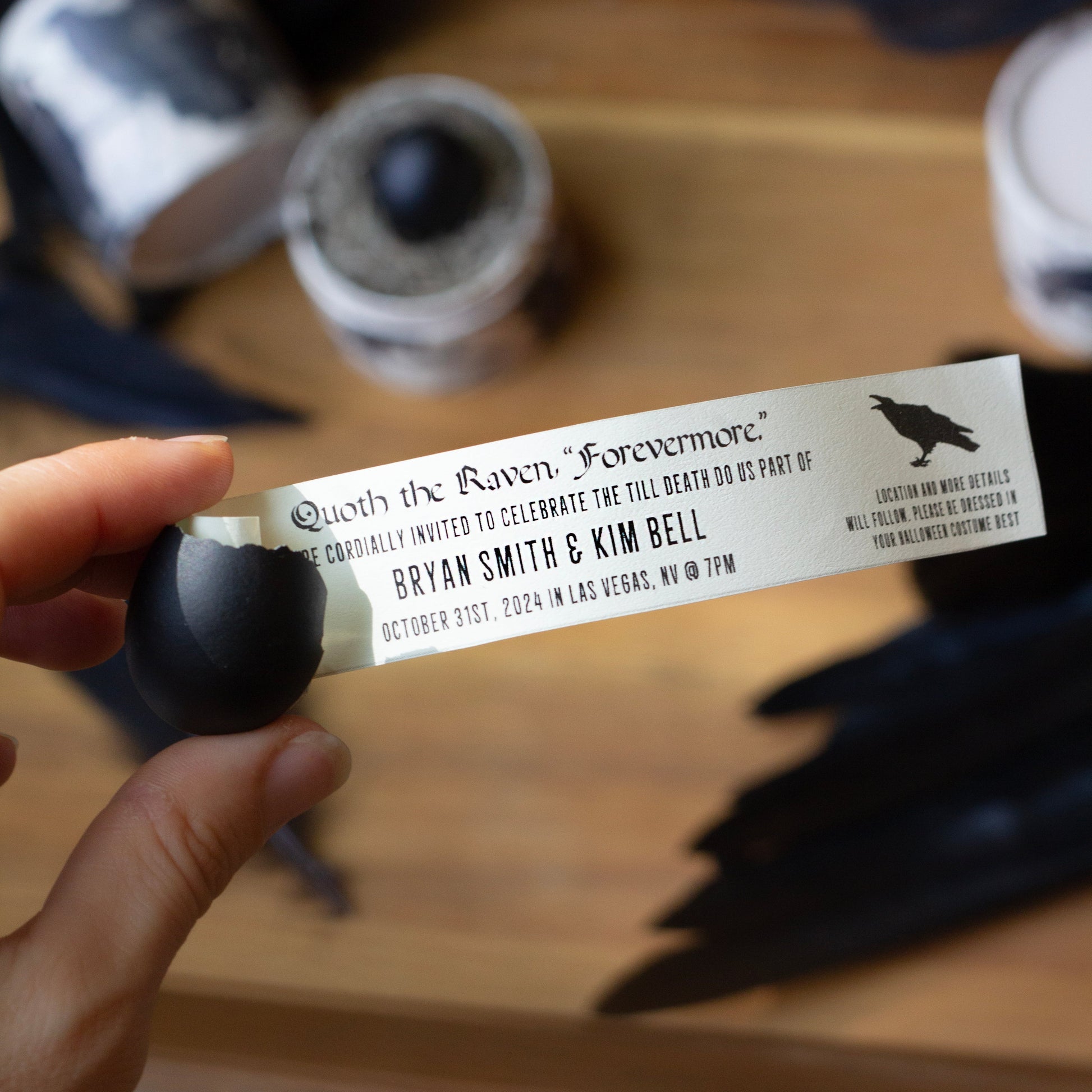 A hand holds a small printed invitation card with black text and a black raven silhouette, surrounded by dark feathers and small glass containers on a wooden surface.