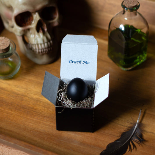 Black egg in a box labeled 'Crack Me' on a wooden surface with a skull and bottles in the background.
