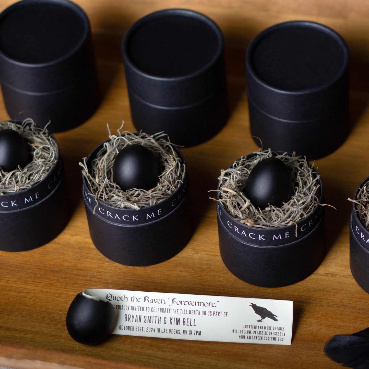 Black egg invitations in black cylindrical tubes with a black band reading 'CRACK ME' on a wooden surface, alongside a printed invitation coming out of a cracked black egg.