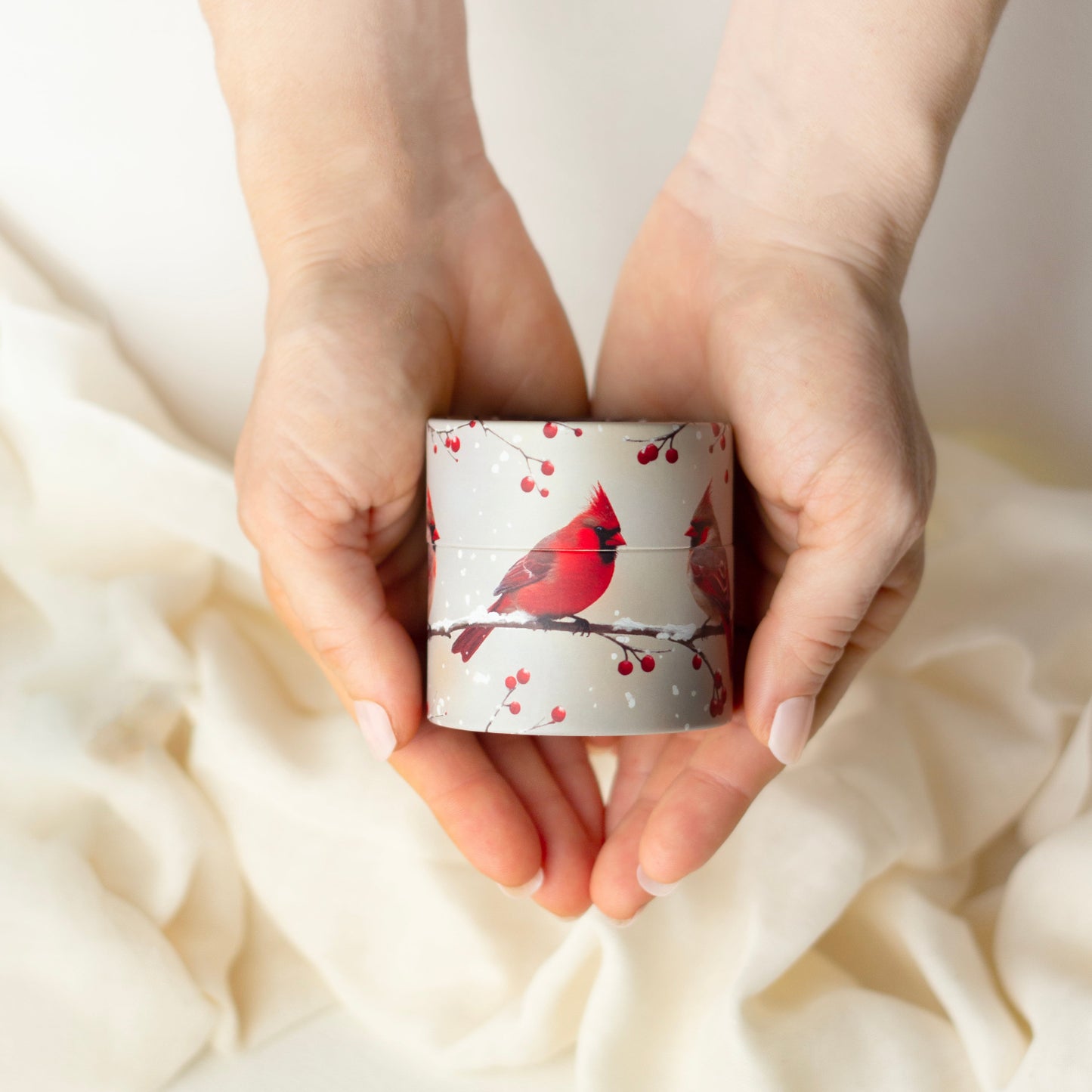Hands holding a small cylindrical container with a red cardinal design on a soft beige fabric background