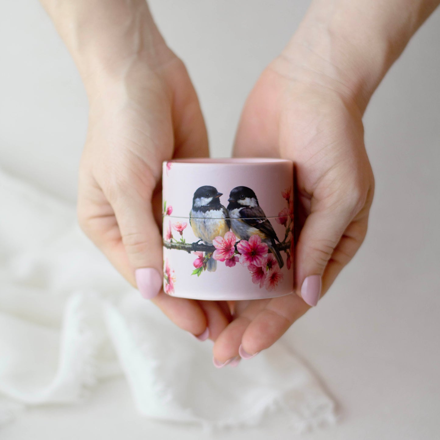 Hands holding a small pink container with bird and cherry blossom design on a light background