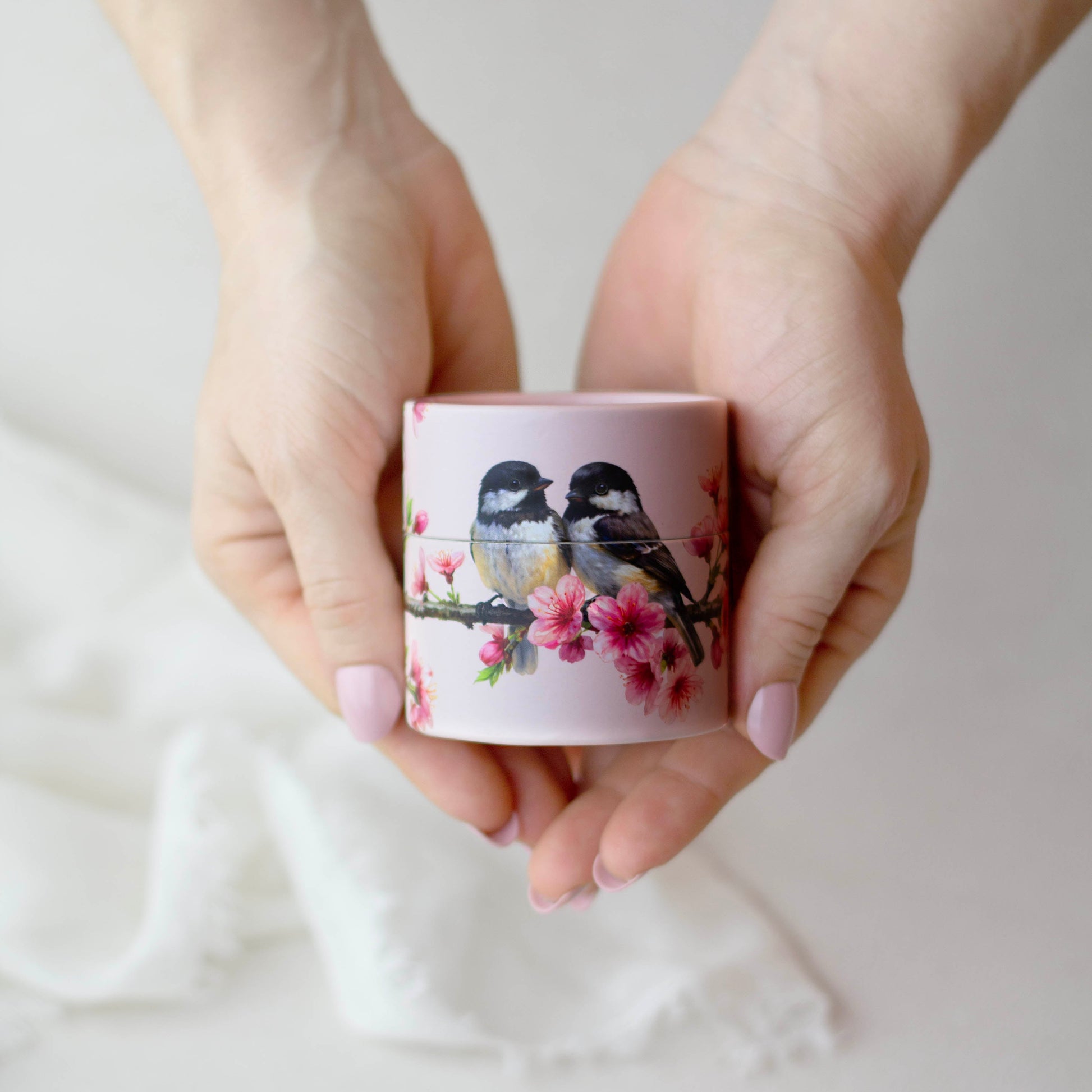 Hands holding a small pink container with bird and cherry blossom design on a light background