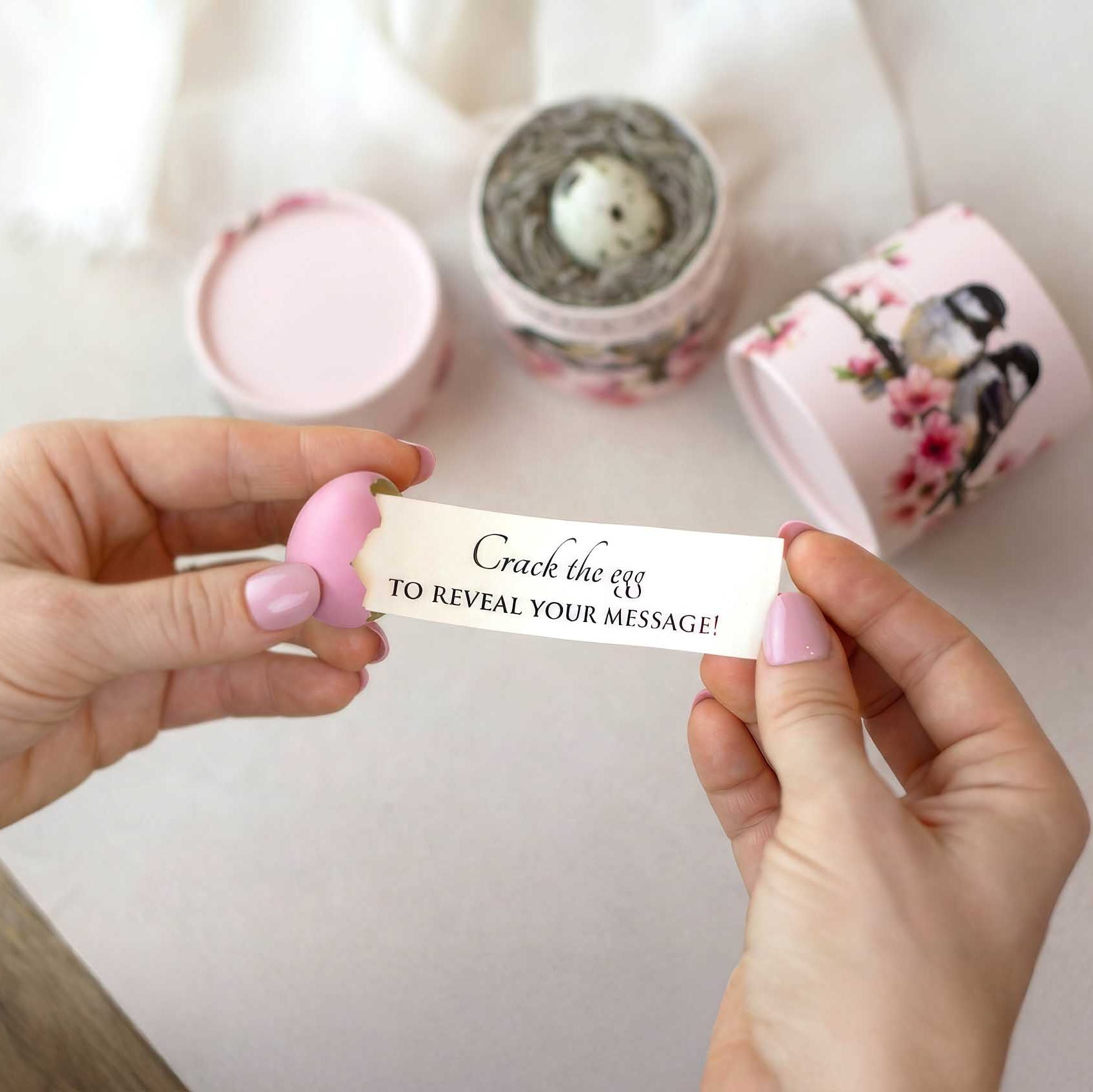 A person's hands hold a small pink egg that is cracked white message being pulled out of it that reads "crack the egg to reveal your message". Pink cylindrical containers with floral and chickadee design sits in the background on a light surface.