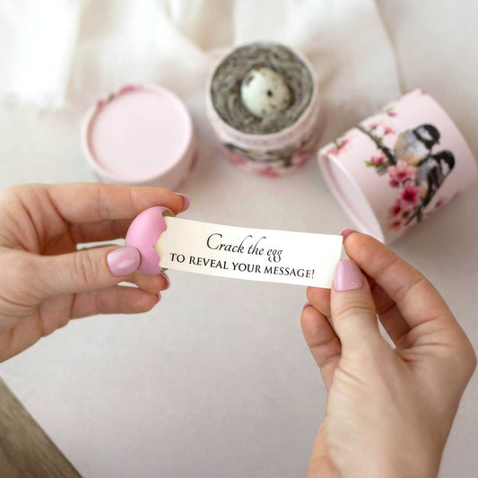 A person's hands hold a small pink egg that is cracked white message being pulled out of it that reads "crack the egg to reveal your message". Pink cylindrical containers with floral and chickadee design sits in the background on a light surface.