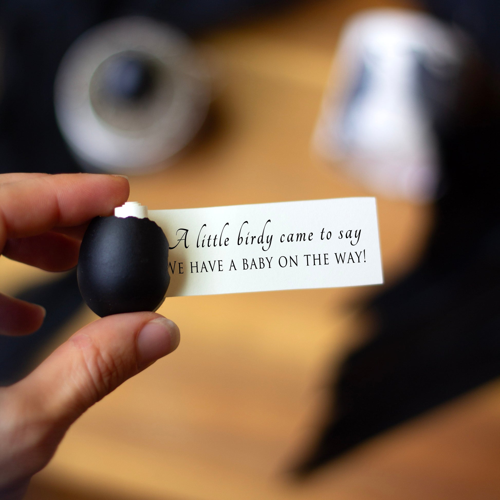 A small black egg with a card reading 'A little birdy came to say we have a baby on the way!' held by a hand.