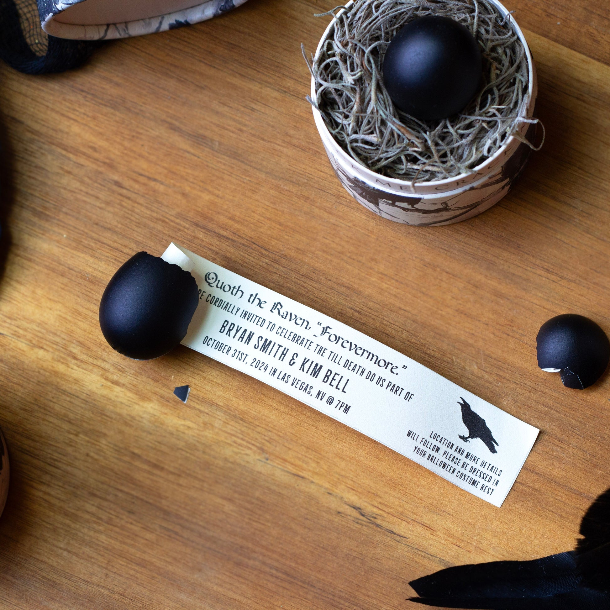 Three matte black eggs arranged on a wooden tabletop with decorative black feathers and small nest-like containers. A printed invitation card lies near the center, and one egg is resting inside a little nest.