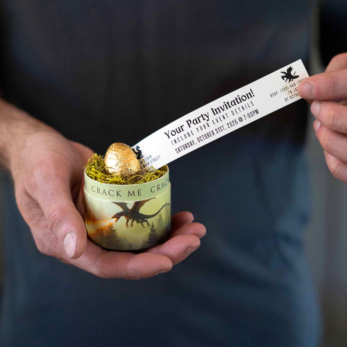 A person holds a small decorative box with dragons on it and a gold dragon egg inside, while sliding a printed paper invitation card out of the cracked egg. The card reads 'Your Party Invitation!' and features a dragon graphic.