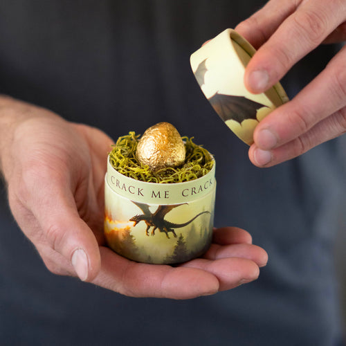 Person holding a small container with a chocolate egg labeled 'Crack Me' against a dark background.