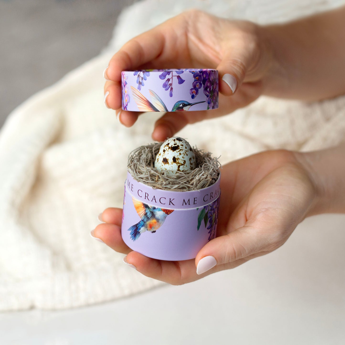 Hand holding a small container with a decorative lid featuring birds and flowers, containing a small bird's nest with eggs.