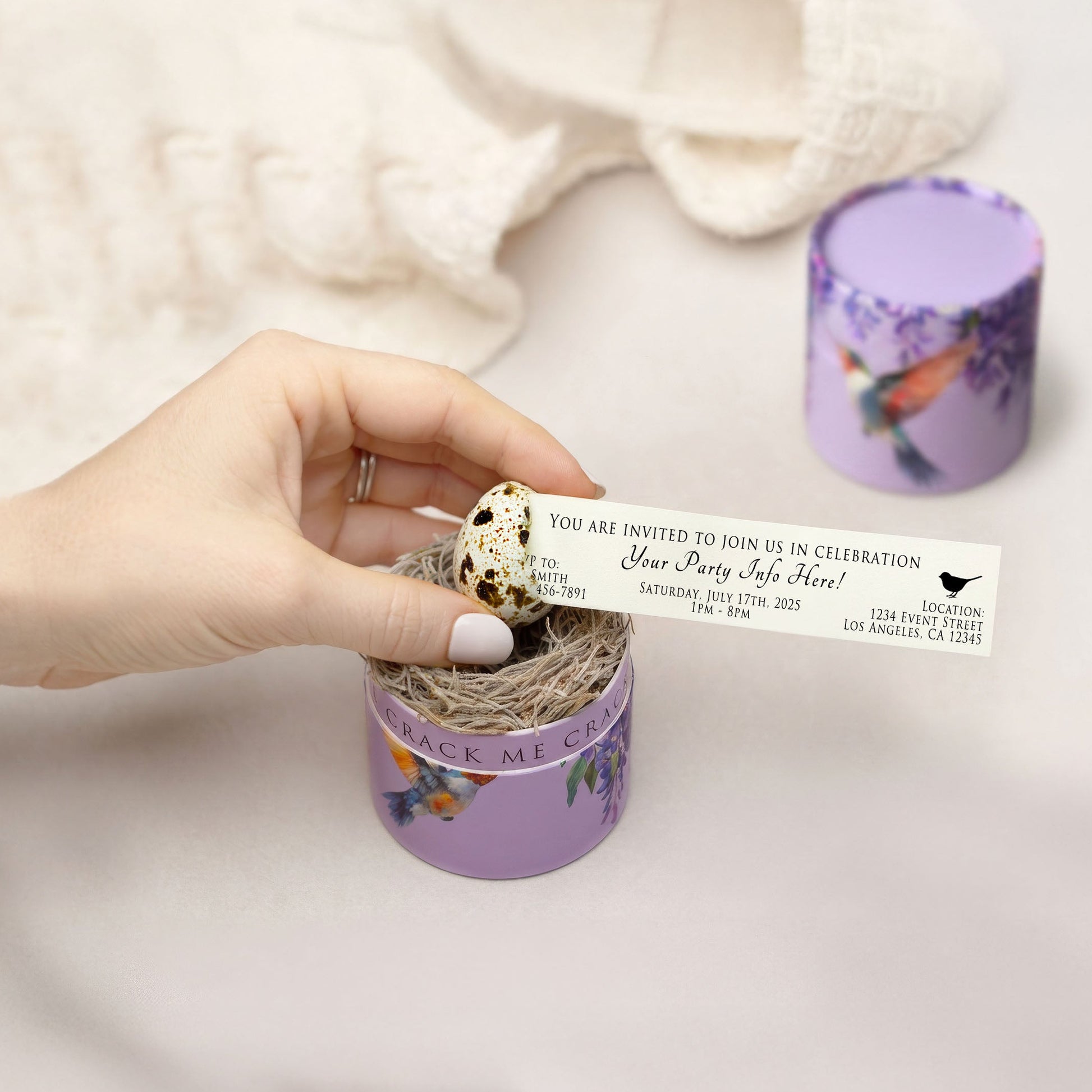 A hand holds a printed invitation card that is being pulled out  of a cracked quail egg shell.  The egg rests upon a moss nest inside a hummingbird themed gift box. 