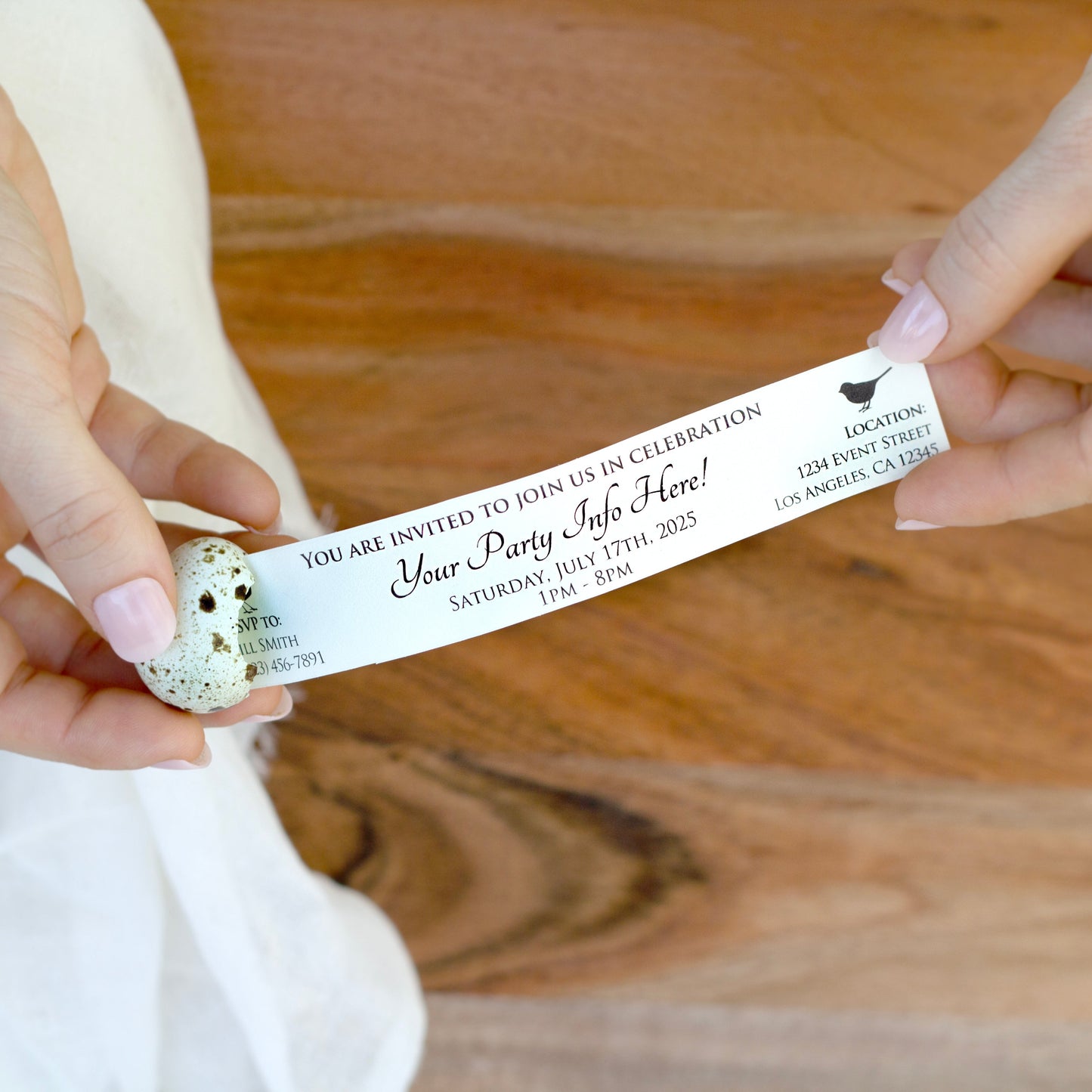 Person holding a party invitation that is being pulled out of the egg on a wooden surface