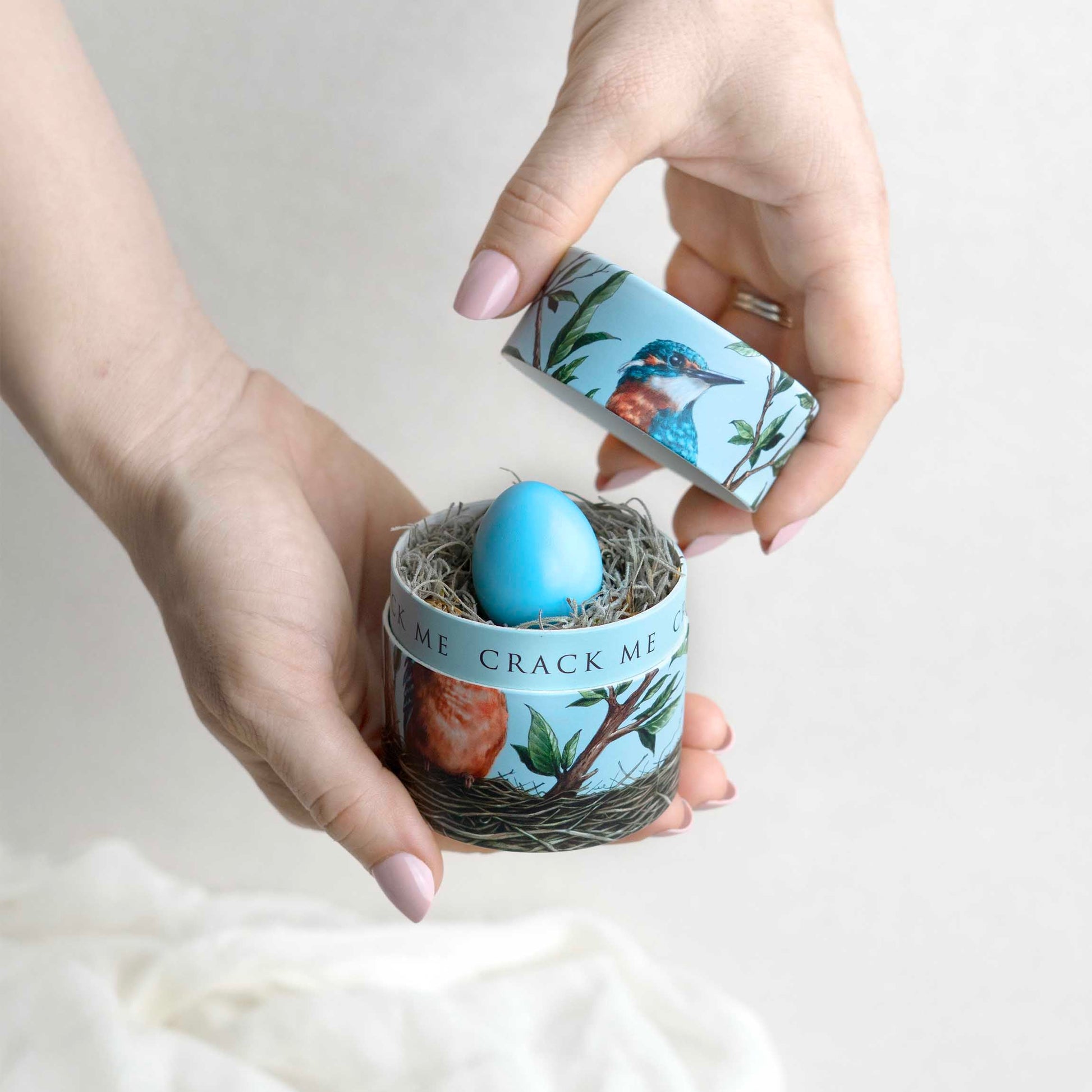Person opening a small container with a blue egg inside, on a light background and kingfisher design on the box