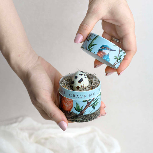 Hands hold a small, blue, kingfisher bird box.  The lid is off and the rim of the box reads "crack me".  Within is a nest with a speckled quail egg. 