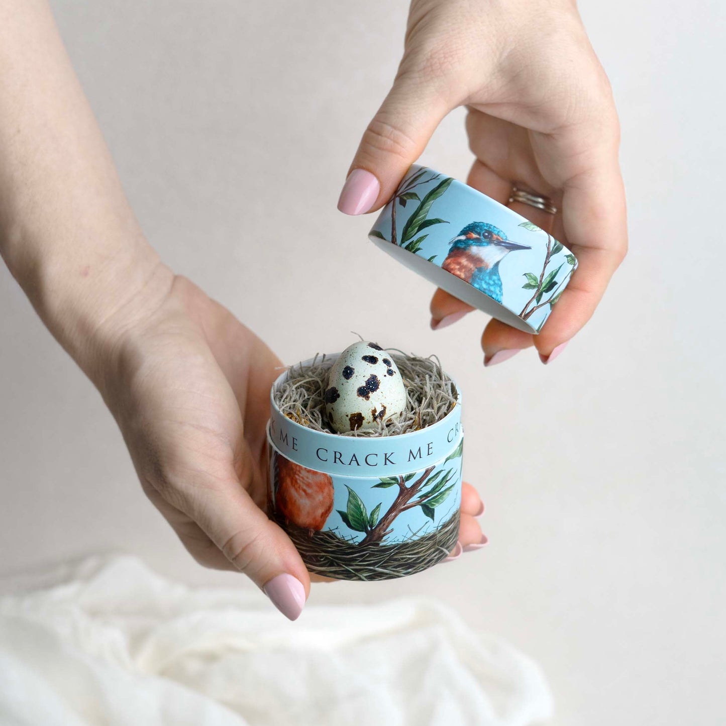 Two hands holding a small container with decorative packaging on a light background and a quail egg inside