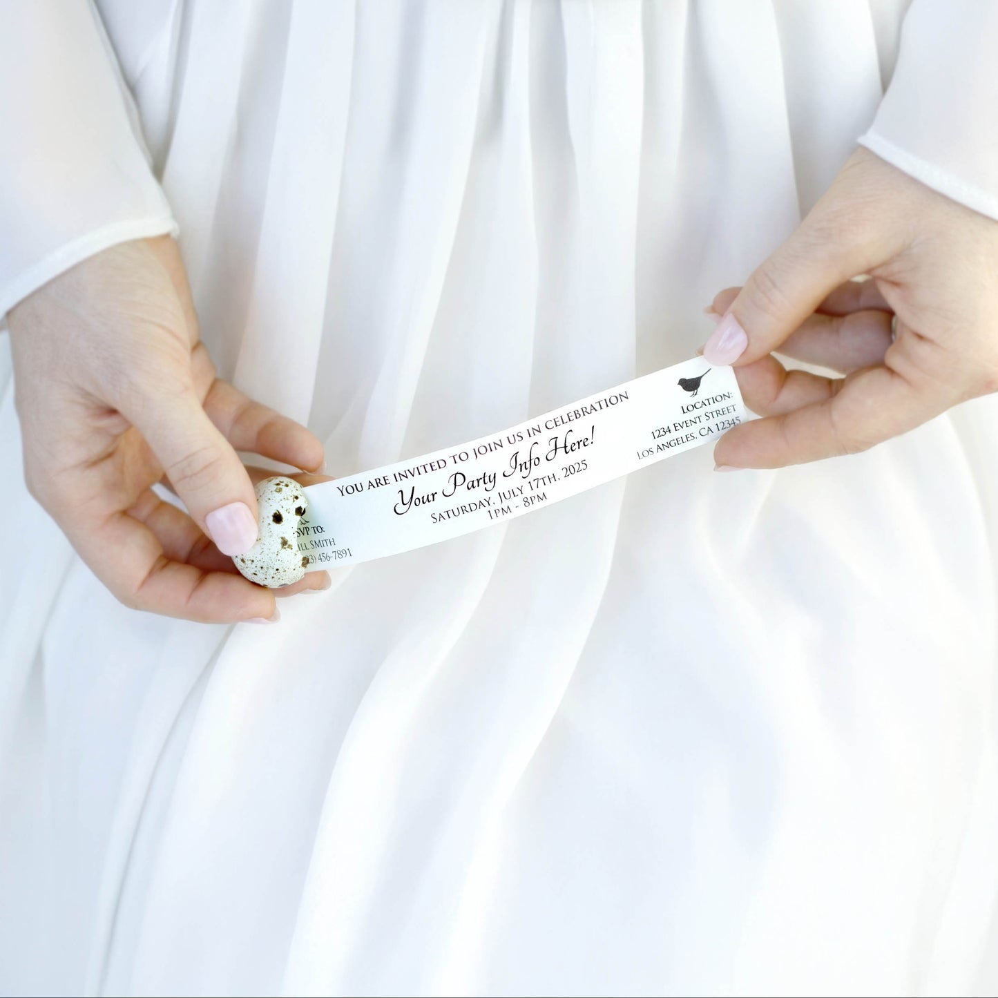 A close-up of a person in a white dress holding a white paper invitation being pulled out of a cracked egg. The invitation is a customizable party invitation.
