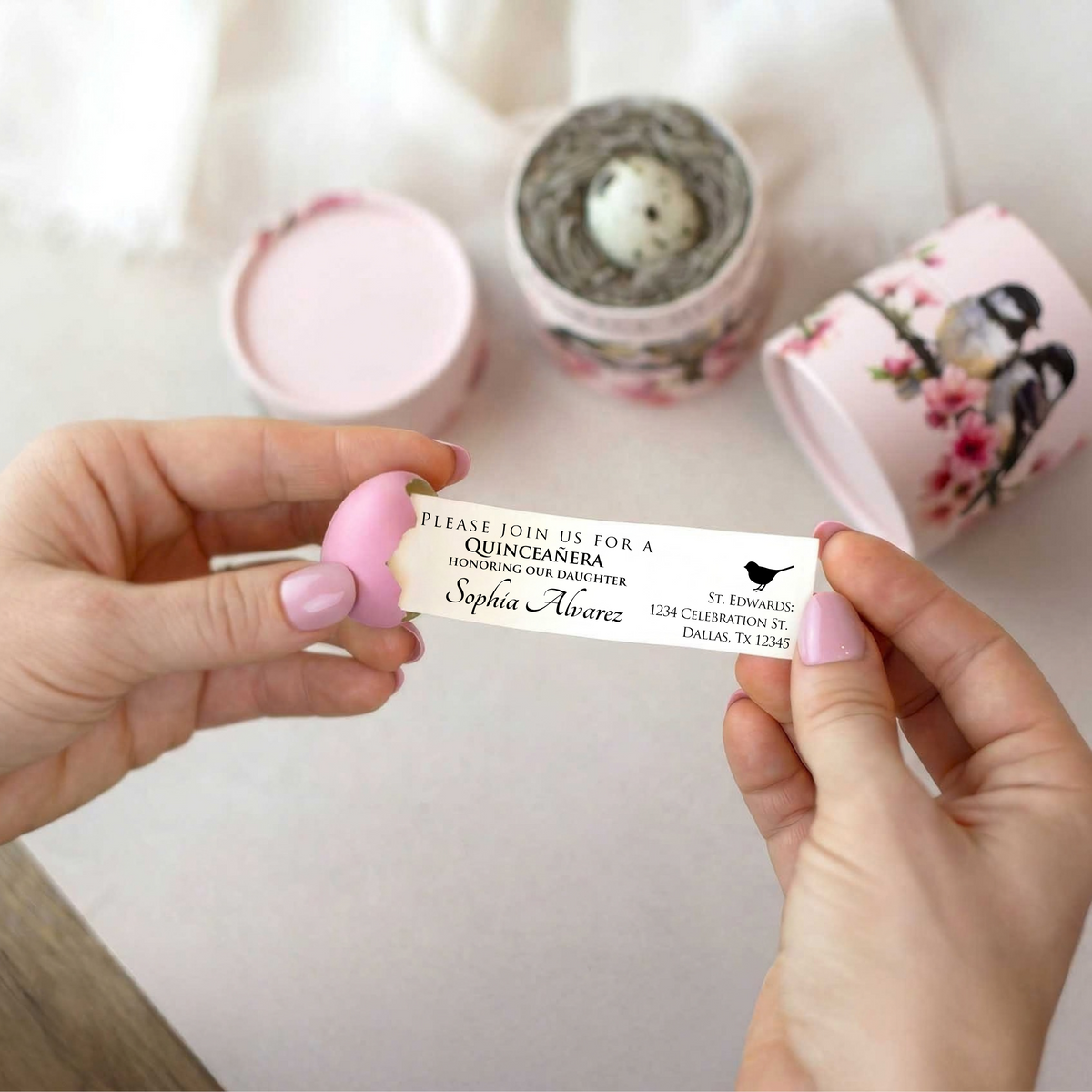 Hands hold a cracked, pink quail egg, with a Quinceanera invitation being pulled out of the egg.