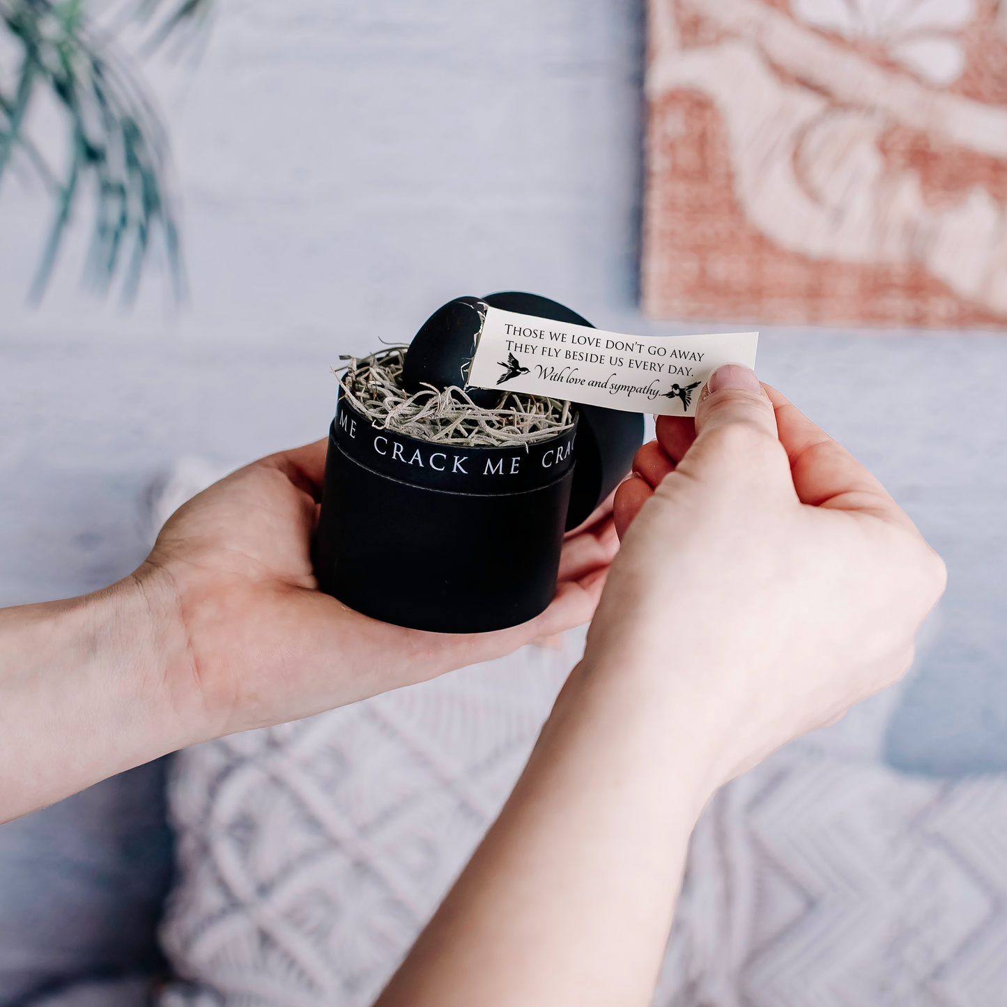 A person's hands holding a black gift box with a nest and a black egg inside. A small custom note is being pulled out of the egg, set against a cozy background with a patterned textile and a plant.
