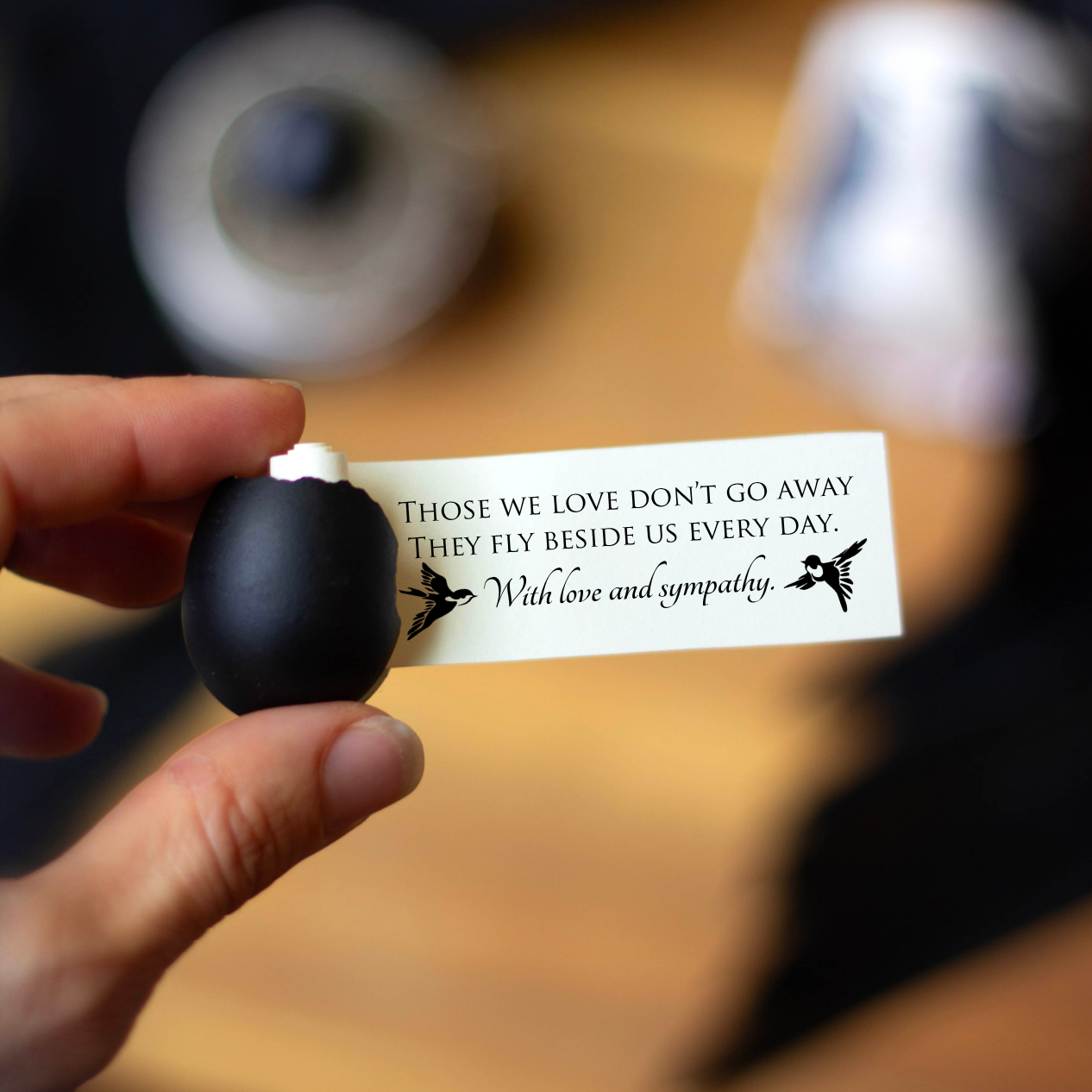 A hand holds a small black egg, the egg is cracked and a message is coming out of it that reads. 'THOSE WE LOVE DON'T GO AWAY THEY FLY BESIDE US EVERY DAY. With love and sympathy,' against a blurred background