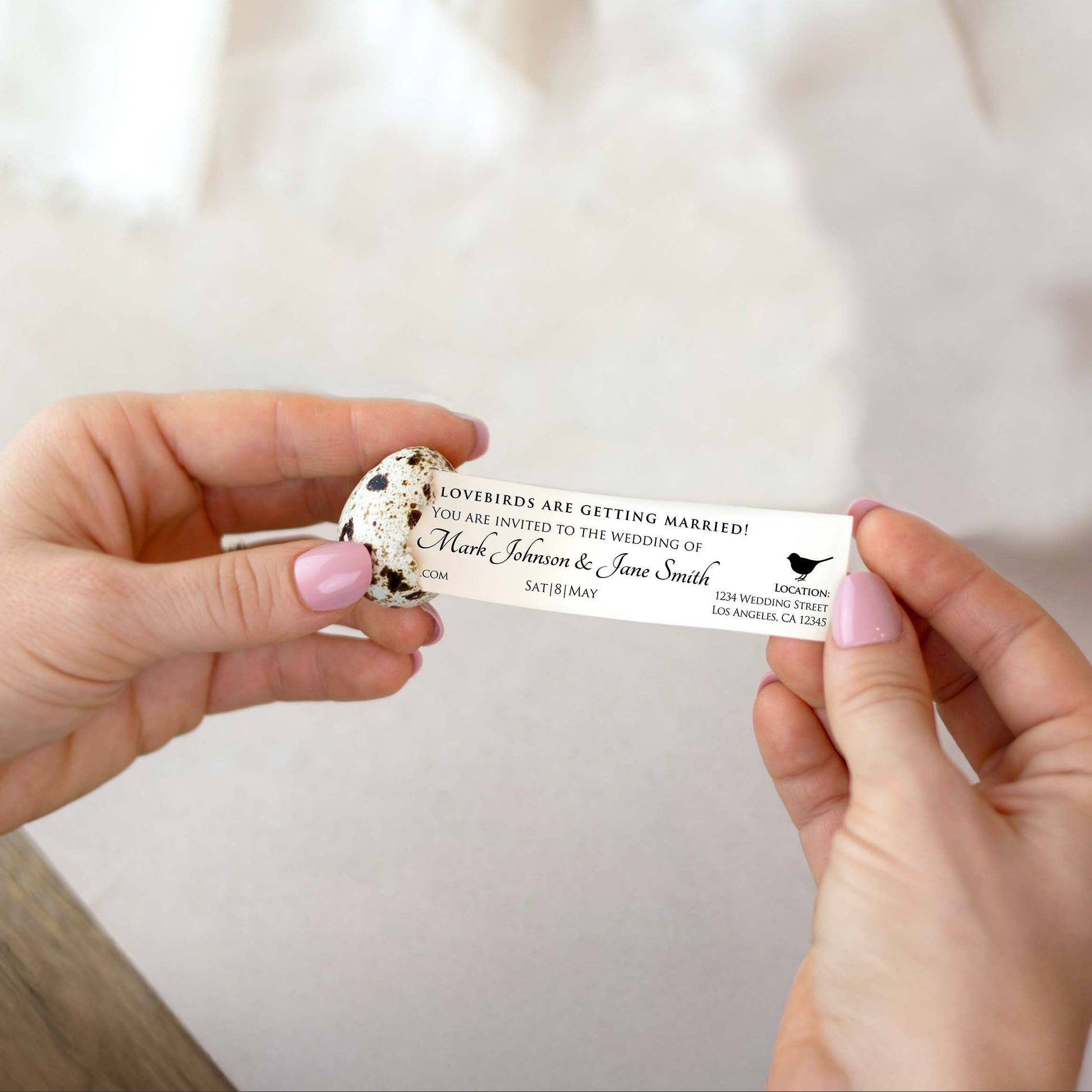 A person pulls a wedding invitation out of a cracked, speckled quail egg shell. The hands have light pink nail polish, and the background is a soft, out-of-focus light neutral surface.