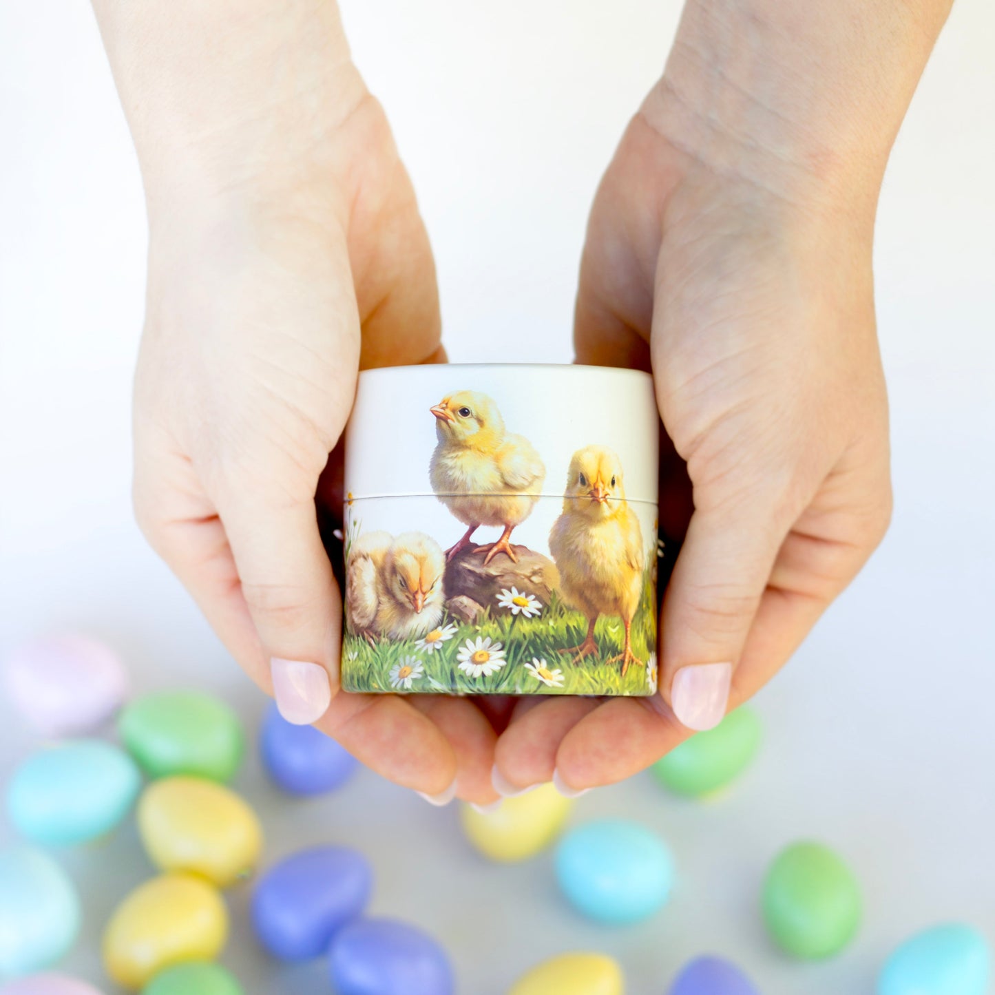 A person's hands holding a small box decorated with a yellow chick illustration, with colorful pastel Easter eggs scattered in the blurred background.