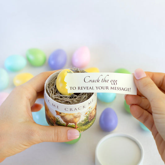 A person holds a small decorative box with a chick image on it and a yellow Easter egg on top. A white paper slip reading 'Crack the egg TO REVEAL YOUR MESSAGE!' is pulled out. of the egg