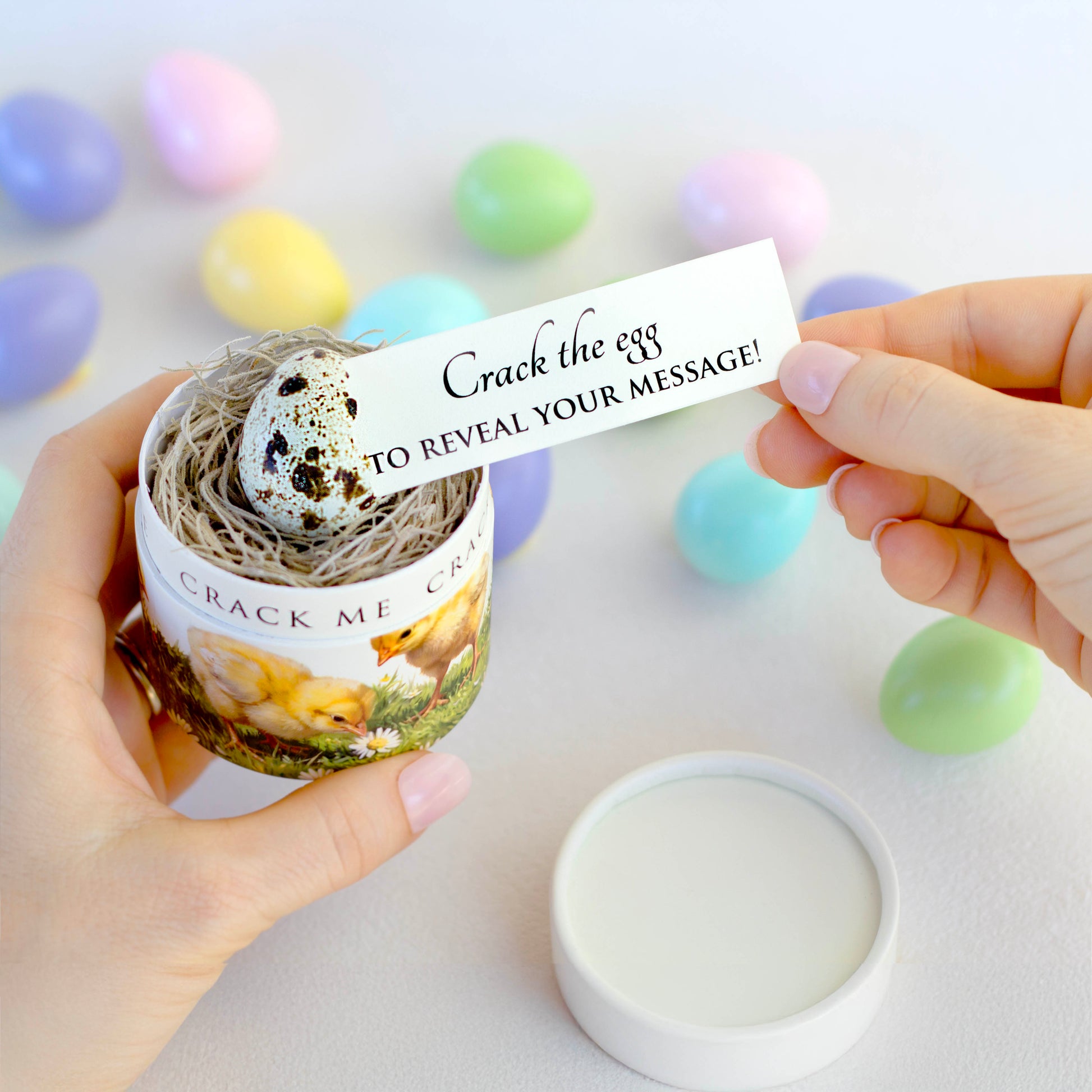 A person holds a small decorative container with a speckled egg inside. The egg is cracked and a paper message is being pulled out, that reads 'Crack the egg TO REVEAL YOUR MESSAGE!'. Colorful pastel Easter eggs are scattered in the background on a light surface.