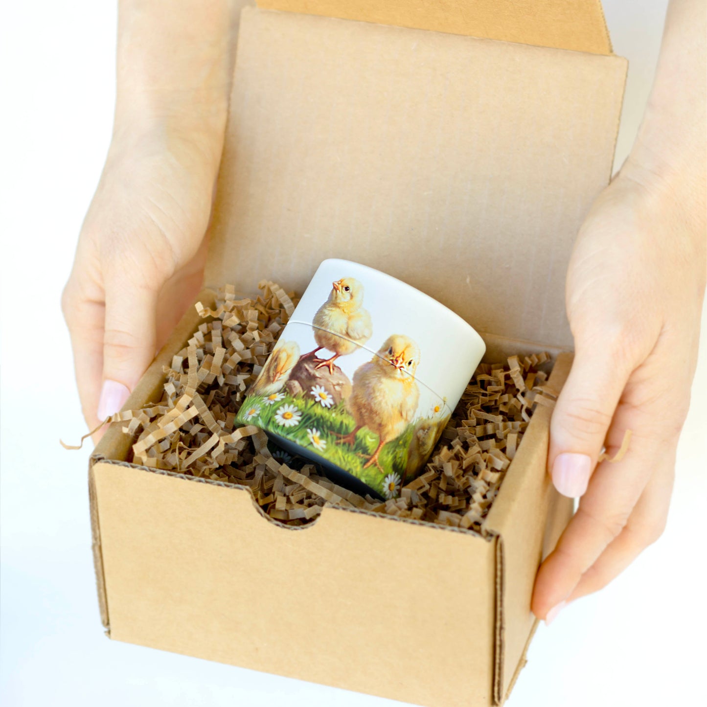 A person's hands holding an open brown cardboard gift box filled with shredded paper and a small gift box featuring two yellow chicks.