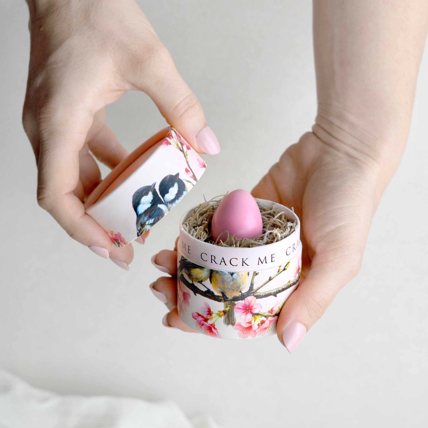 A person's hands holding a small floral- an chickadee patterned container with a pink egg-inside, against a soft white background.