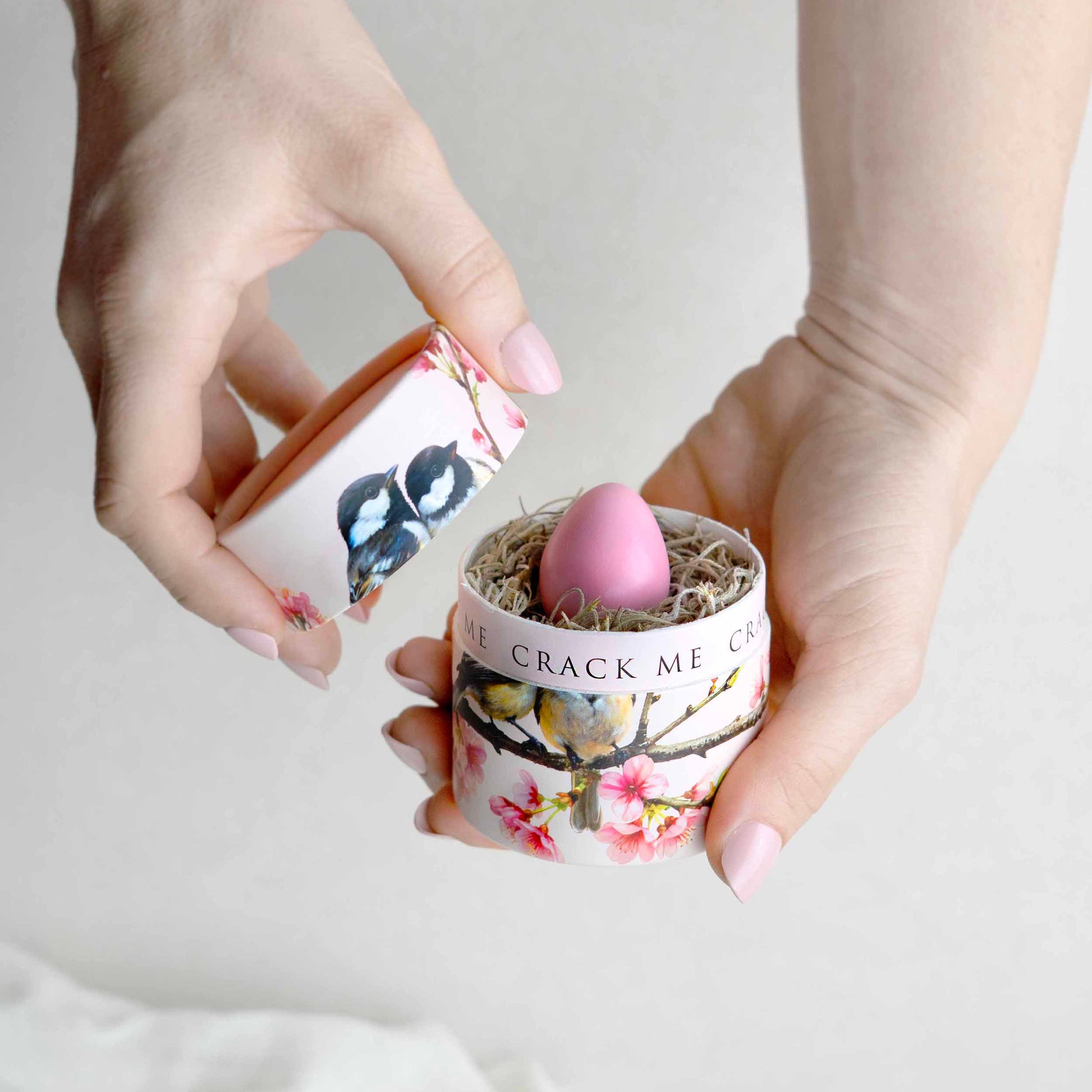 A person's hands holding a small floral- an chickadee patterned container with a pink egg-inside, against a soft white background.