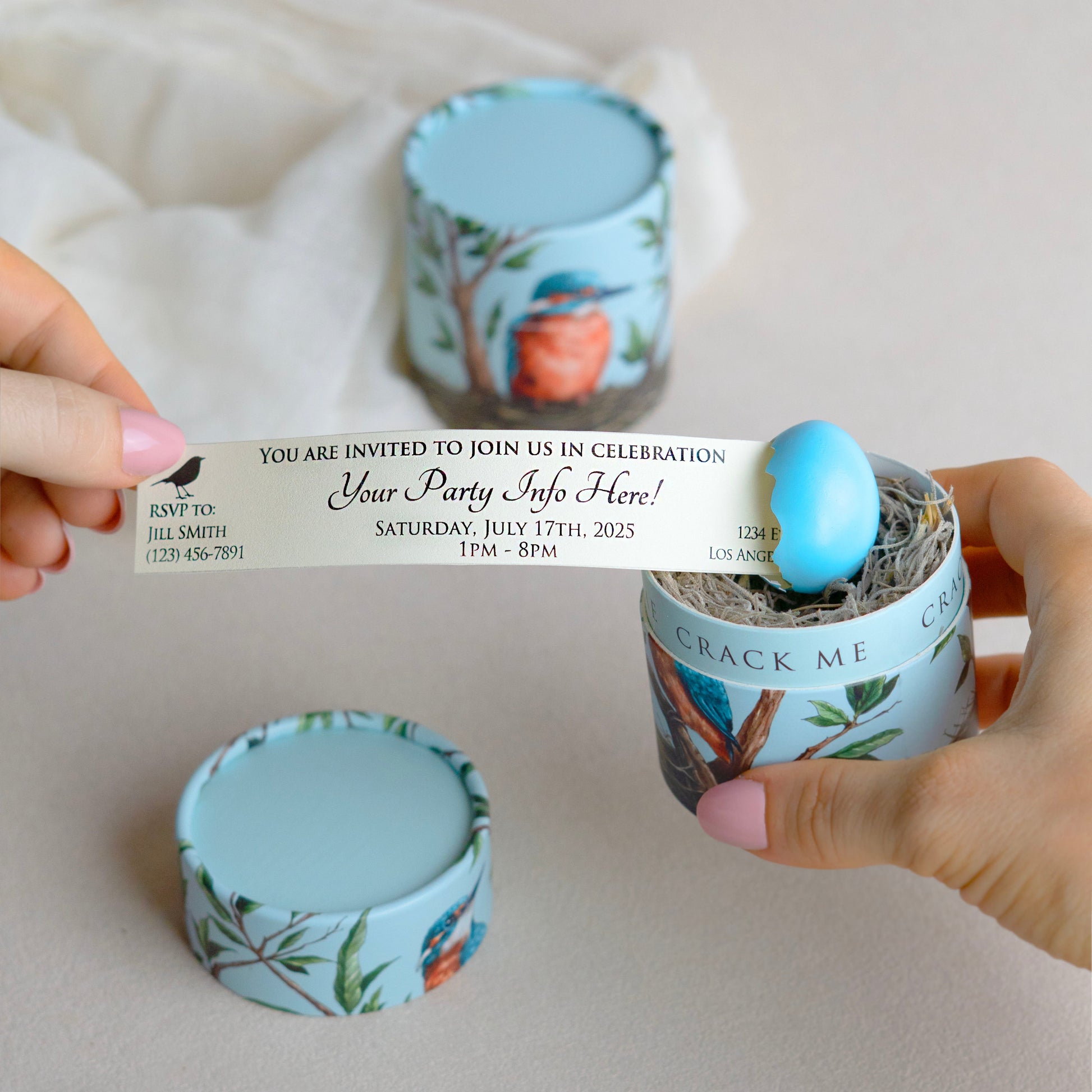 A person's hands hold a printed invitation that is being pulled out of a small blue egg. The egg sits within a moss nest inside of a gift box with a botanical and bird illustration.
