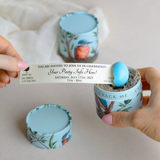 A person's hands hold a printed invitation that is being pulled out of a small blue egg. The egg sits within a moss nest inside of a gift box with a botanical and bird illustration.